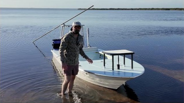 Capt. Rick standing knee-deep beside the East Cape Evo 17 skiff in the lagoon shallows, poling platform behind him